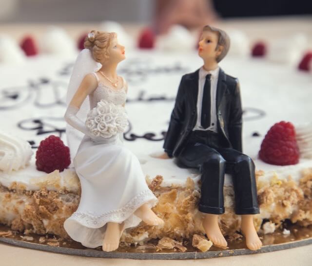 Picture of two traditional wedding cake figurines of a bride and groom sitting on the edge of the wedding cake talking.
