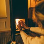 Photo of a woman sitting in front of a wood stove warming her hands by the fire.