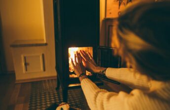 Photo of a woman sitting in front of a wood stove warming her hands by the fire.