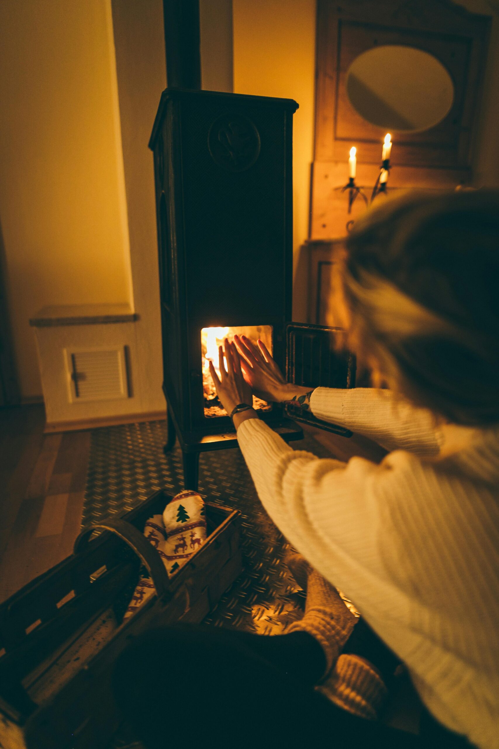 Photo of a woman sitting in front of a wood stove warming her hands by the fire.